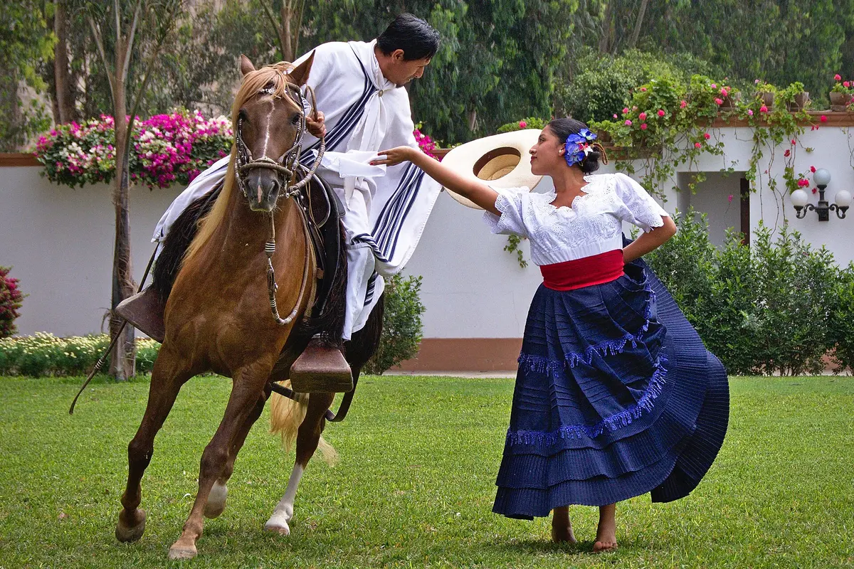 A barefoot dancer in a blue skirt and white blouse extends her arm toward a mounted chalan in a white poncho on a hacienda lawn